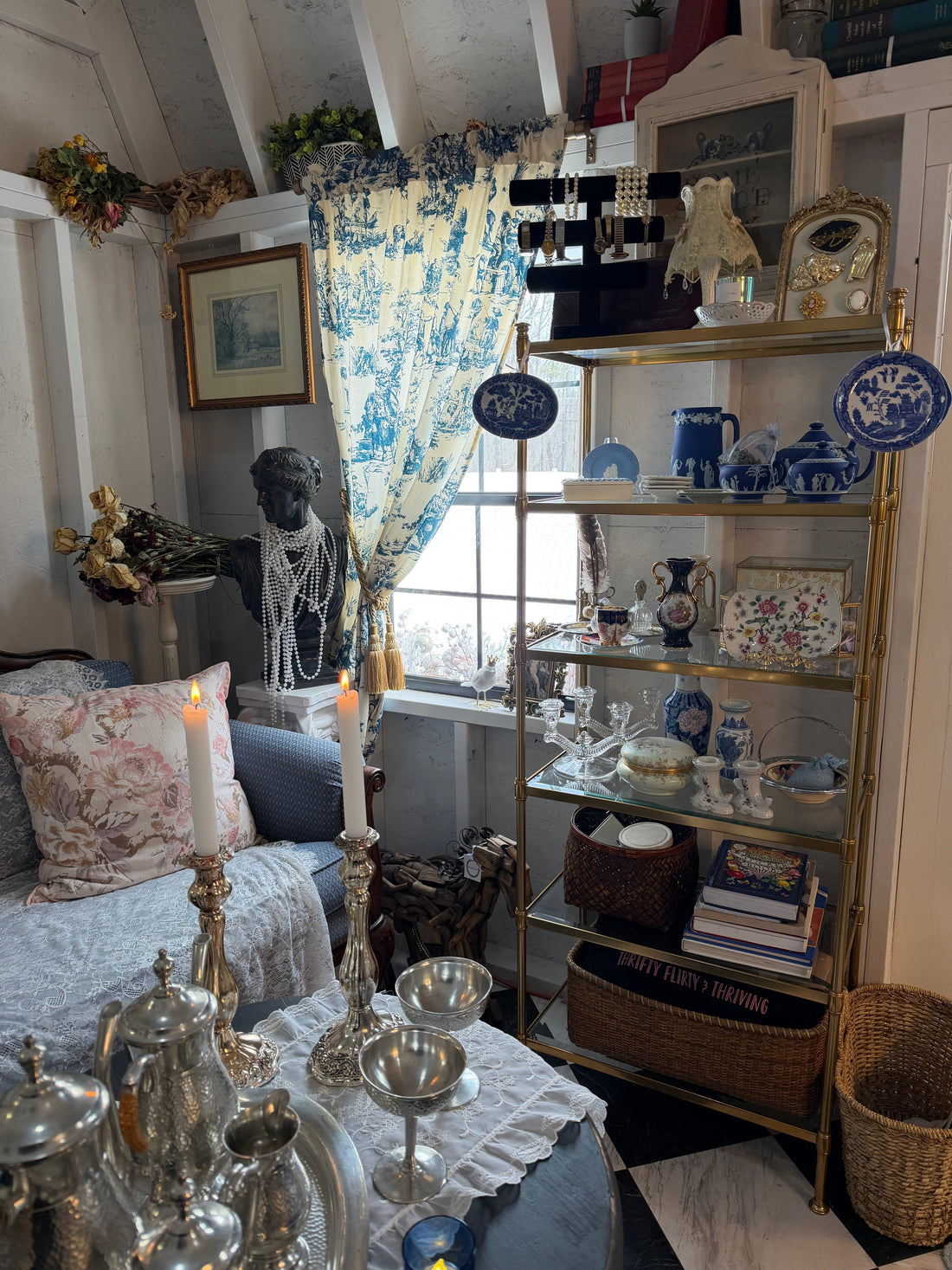She Shed studio styled with European-inspired vintage decor featuring blue and white china, brass shelving, antique glassware, a silver tea set, and a classical bust with pearl necklaces beside blue toile curtains and a cozy chair.